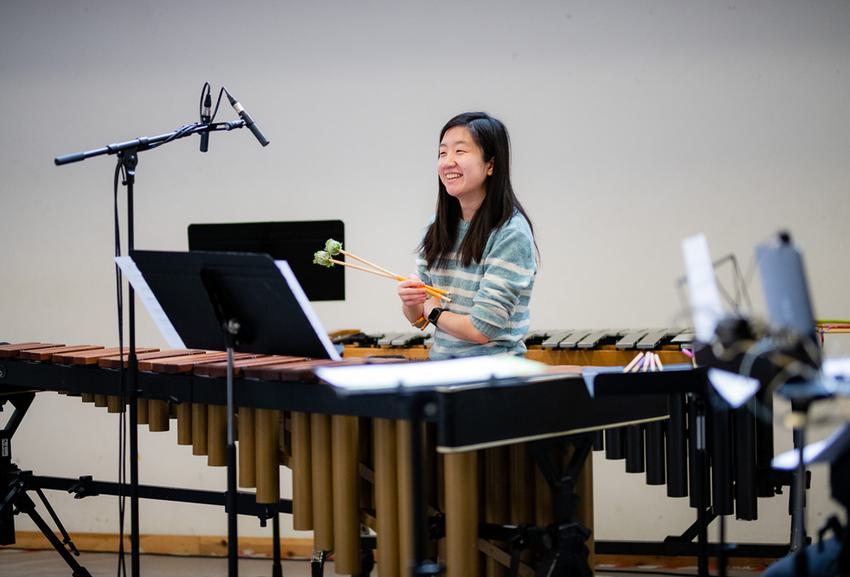 A young percussionist with long dark hair and medium light skin tone stands in from of a marimba, smiling and holding her mallets