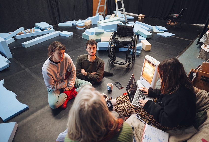 Four artists sit on the floor and on a sofa in a brightly-lit rehearsal space, deep in discussion as one takes notes on a laptop. Foam blocks are strewn across the floor behind them.