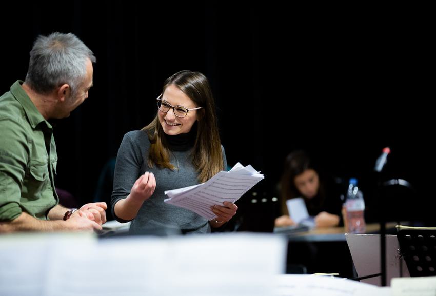 A young woman with long brown hair and glasses with light skin tone gesticulates as she discusses sheet music with a man with gray hair who is facing away from the camera.