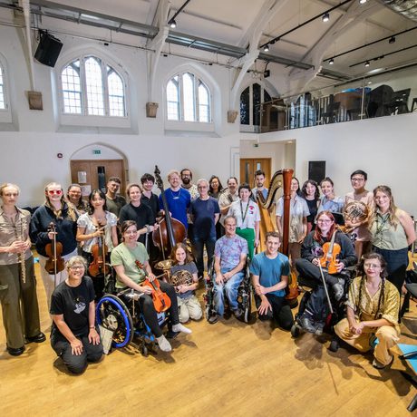 A group of musicians and composers are gathered together, facing the camera and smiling. Many of them are holding their instruments and they are standing in a brightly-lit rehearsal space.