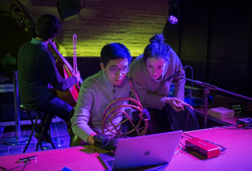 Two musicians, one holding a natural horn and wearing a pair of gloves covered with LEDs and wires, look intently at a laptop together.