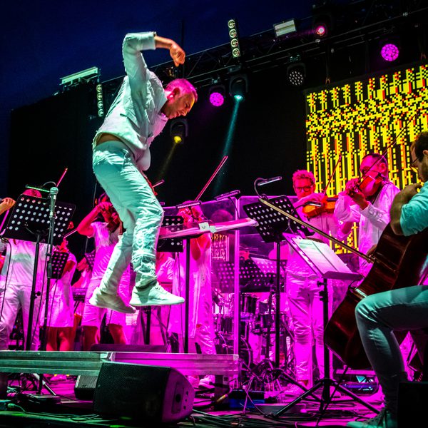 Charles Hazlewood, a man with light skin tone wearing a white suit, leaps in the air as he conducts the string section of Paraorchestra on stage at WOMAD festival. Behind the musicians, spotlights and an LED screen cast vivid purple & yellow light across the scene.