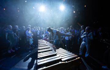 An audience watching three musicians perform in Paraorchestra's 'The Nature of Why' at the Southbank Centre in London. One performer is playing the marimba while held in the air by another performer. The room is dark with bright white lights illuminating the performance.