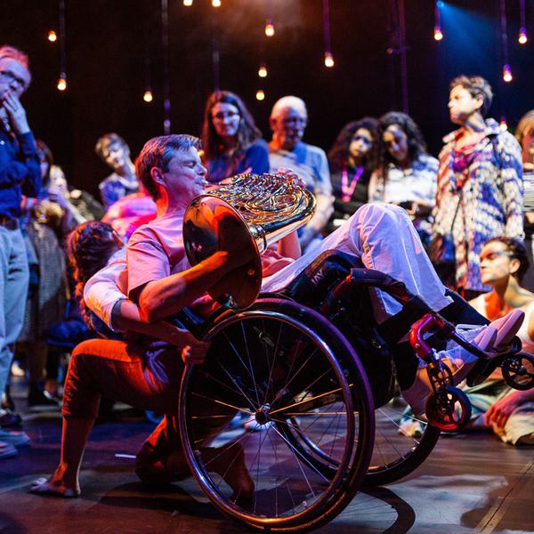 A musician and a dancer are performing with audience members surrounding them in a circle, watching the performance intently. The musician is playing a horn, wearing a pink t-shirt, blue jeans and is a wheelchair user. Their wheelchair is balancing on its back wheels, lifted into the air and being supported by the dancer, who is faced towards the audience. The lighting is atmospheric and the performers are illuminated in contrasting blue and pink hues.