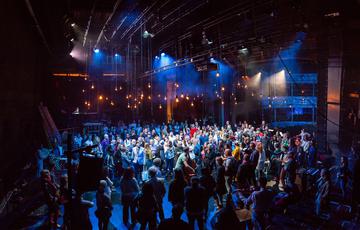 Inside a large theatre, audience and performers mingle together on a stage lit by dozens of filament bulbs suspended from the ceiling. Amongst the crowd a harp, double bass, and other string instruments are visible.