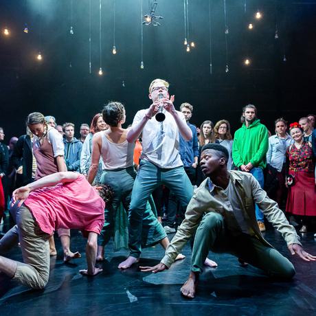 Four dancers and one musician are performing in a brightly lit room, with audience members surrounding them in an immersive and intimate setting. The musician is playing the clarinet and the dancers are in motion.