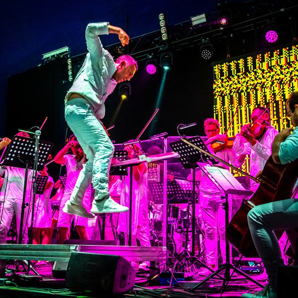 Charles Hazlewood, a man with light skin tone wearing a white suit, leaps in the air as he conducts the string section of Paraorchestra on stage at WOMAD festival. Behind the musicians, spotlights and an LED screen cast vivid purple & yellow light across the scene.
