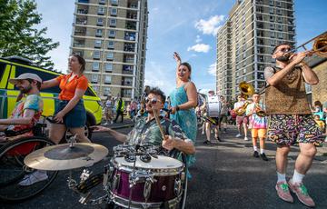 A male in sunglasses plays a drum mounted in front of his wheelchair, flanked by a dancer and trombonist. They are followed by other musicians, playing brass and percussion, all on parade down the street towards us. There are bright blue skies seen in the background and they are in an urban setting with two high rise apartment blocks and an emergency vehicle in the background.