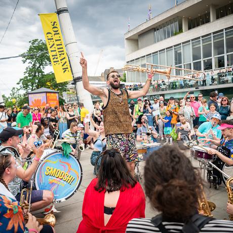 A huge crowd gathers in a circle around musicians crouched playing percussion, brass, and woodwind. They are performing outside the Southbank Centre, which can be seen to the right. In the centre, a man wearing sunglasses and a leopard-print top, holds a trombone. His arms are outstretched and he points to the sky, his mouth open with joy as he encourages the crowd.