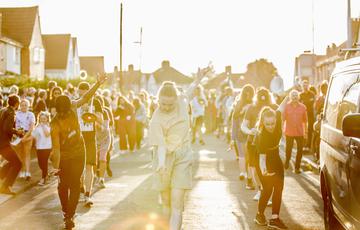 Musicians and dancers parade through a suburban street in Bristol on a sunny Summer's day. The sun is casting a golden light across the scene and the street is lined with spectators.