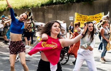 A trio of colourfully-dressed dancers perform in a sunlit car park, with musicians and more dancers visible behind them.