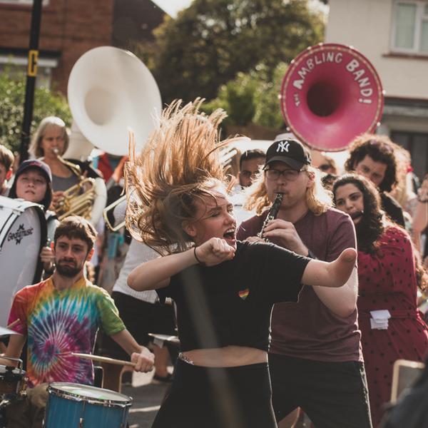 A female dancer flicks her hair and smiles energetically as she performs amidst a crowd of brass and woodwind players on a suburban street.
