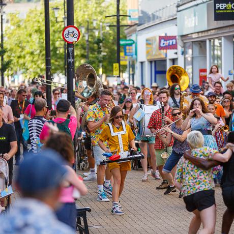 Paraorchestra musicians performing on the streets of Weston-Super-Mare. They are walking down a busy high street and are playing a wide range of instruments including a flute, clarinet and a drum. There is a large audience who are also walking down the busy street. It is daytime and the image is brightly lit.