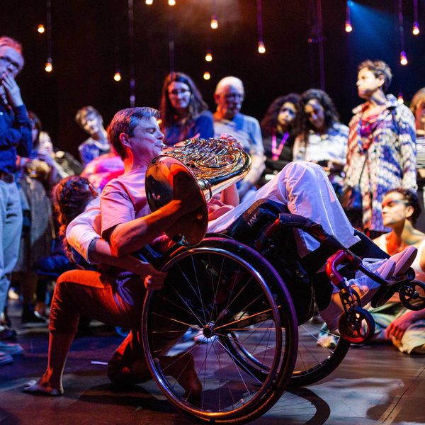 A musician and a dancer are performing with audience members surrounding them in a circle, watching the performance intently. The musician is playing a horn, wearing a pink t-shirt, blue jeans and is a wheelchair user. Their wheelchair is balancing on its back wheels, lifted into the air and being supported by the dancer, who is faced towards the audience. The lighting is atmospheric and the performers are illuminated in contrasting blue and pink hues.