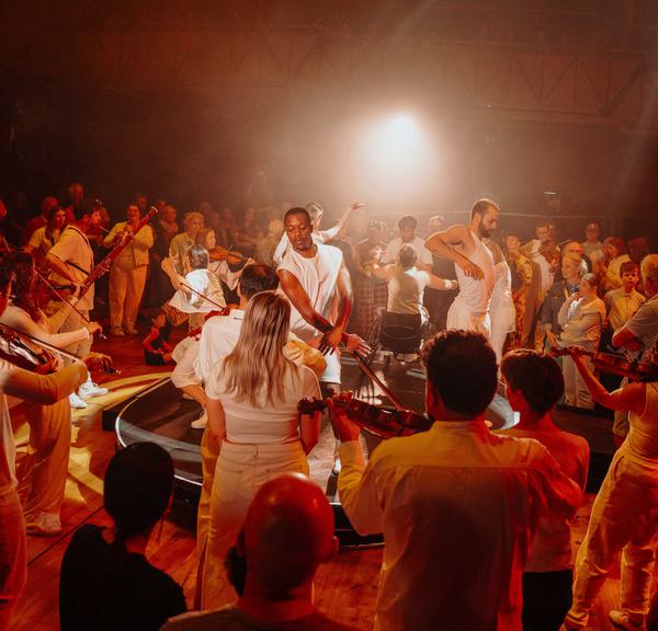 A group of Paraorchestra musicians and dancers are standing in a circle, facing both inwards and outwards. They are all wearing white with a warm orange light glowing above them, illuminating the performers in hues of red and orange. A large audience is visible in the foreground and background, intently watching the performance.
