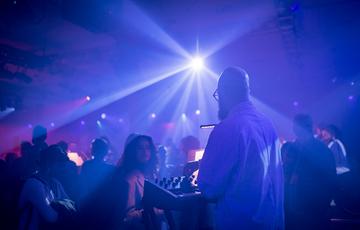 A synth player with glasses, a beard and light skin tone wearing all white performs to a small audience
