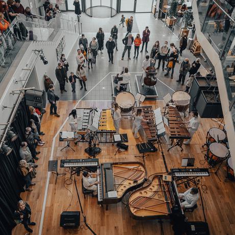 A top-down view showing around a dozen Paraorchestra musicians playing percussion and piano. Audience members lean over to look down from staircases and mezzanines above.