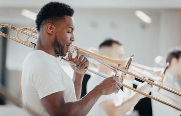 A male trombone player with a beard, short hair, and medium dark skin tone performs in a brightly-lit atrium.