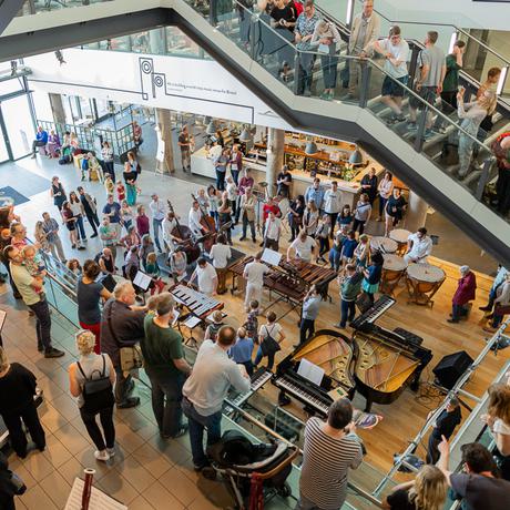 Around a dozen Paraorhcstra musicians play piano and percussion on the floor a large atrium lit with sunlight. Surrounding them on the floor and looking on from above are around one hundred audience members.