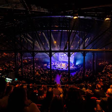 A wide-angle shot of the Roundhouse in London. There is a large seated audience watching Death Songbook be performed. The stage is illuminated in blue hues while the audience are illuminated in warm orange lighting.