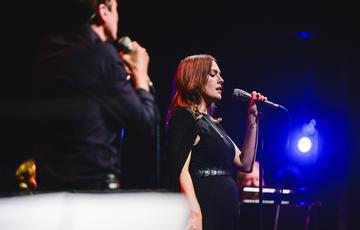 Gwenno, a woman with shoulder-length brown hair, wearing a black cape, black top and black jeans is also holding a microphone and singing. In the foreground of the image and slightly out-of-focus, Brett Anderson, a man with fair-skin and short dark hair wearing a black shirt and black jeans, is holding a microphone and also singing on stage. The stage is dark and illuminated in blue hues, while the two performers are brightly lit in white spotlights.