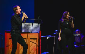 Brett Anderson, a man with fair-skin and short dark hair wearing a black shirt and black jeans, is holding a microphone and singing on stage. Gwenno, a woman with shoulder-length brown hair, wearing a black cape, black top and black jeans is also holding a microphone and singing. The stage is dark and illuminated in blue hues, while the two performers are brightly lit in white spotlights.