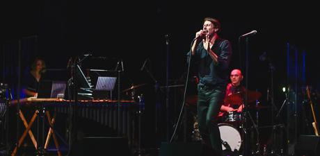 Brett Anderson, a man with fair-skin and short dark hair wearing a black shirt and black jeans, is holding a microphone and singing on stage. Behind him, a drummer and a xylophonist are also performing on their respective instruments. The stage is dark and Brett is illuminated by a white spotlight.