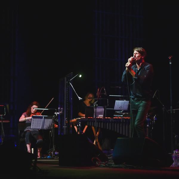 Brett Anderson, a man with light skin a short, dark hair is wearing a black shirt and trousers. He is performing on stage, singing into a microphone. Four musicians are performing with him, including a double bassist, a cellist and a violinist. The stage is dark and the backdrop is black.