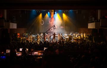 Paraorchestra perform on a large stage illuminated by blue, orange and yellow spotlights. In the foreground the silhouetted heads of a large audience are visible, along with the glowing controls of the sound desk.