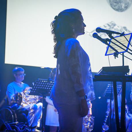 A female singer with red hair a light skin tone wearing all white stands up straight as she prepares to sing, semi-silhouetted against a large video screen