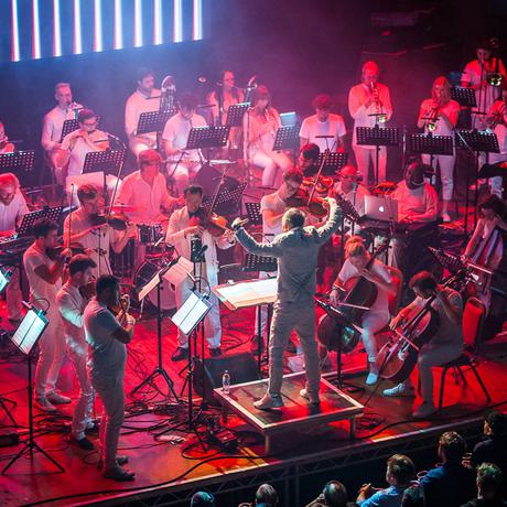 Charles Hazlewood, a man in his fifties wearing a silver suit, faces the Paraorchestra musicians as he conducts, hands held aloft