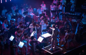 The string section of Paraorchestra, all wearing white, perform as a conductor gestures in front of them on a stage illuminated in purple