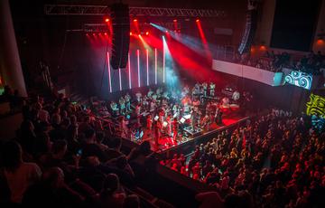 Approximately twenty Paaorchestra musicians perform to a large audience in a concert hall on a stage illuminated with vivid red spotlights