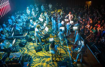Around two dozen Paraorchestra musicians, all wearing white, perform on a stage illuminated in swirling blue-green light.