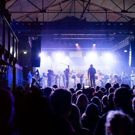 A large crowd watches Paraorchestra players perform on stage, their figures silhouetted in purple light.