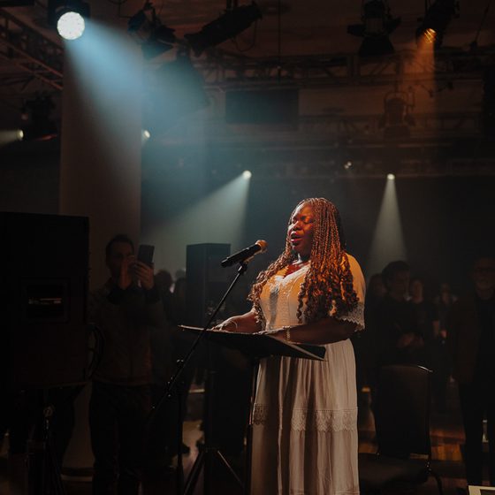 Victoria Oruwari, a woman wearing a white dress with long golden braids and medium ark skin tone, reads braille music as she sings.