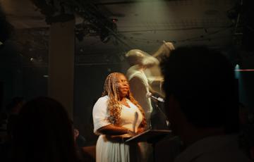 Victoria Oruwari, a woman with long golden hair in braids and medium dark skin tone wearing a white dress reads braile sheet music with her hands as she sings. Behind her a conductor gestures in the air.