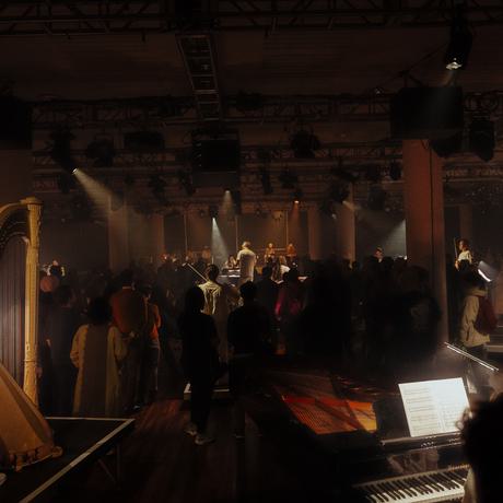 A wide shot of a darkened performance space. Musicians dressed in white including a harpist, pianist, and string section stand on small platforms with the audience silhouetted around them.