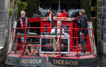 Steve Varden, a Paraorchestra musician smiles as he plays electronic instruments aboard a canal boat whilst it descends in a canal lock with cascading water behind it.