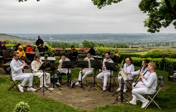 Eight Paraorchestra musicians playing wind instruments perform outdoors. They are all dressed in white and a rolling green country landscape is visible behind them.