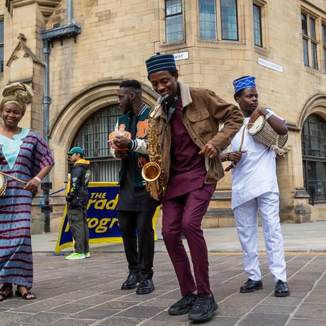 A group of musicians led by Oluwatosin Omotola Ajayi wearing vibrant Yoruba dress play percussion and saxophone outside of Bradford City Hall.