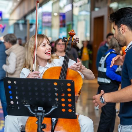 A Paraorchestra cellist seated with her instrument chats with an audience member inside a shopping centre. She has blonde hair and is smiling warmly.