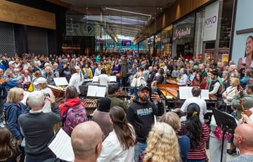 A large crowd of hundreds of people gather around a pianist and percussion section from Paraorchestra inside a large modern shopping centre.