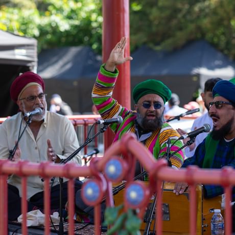 Three members of the Shah-e-Mardan Qawwali group perform in a bandstand on a sunny day. They are seated cross legged on the floor wearing colourful traditional dress, singing an clapping their hands. One member plays a shruti, a box-shaped instrument that produces sound through bellows.