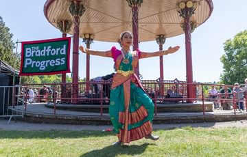 A young female dancer from the Annapurna Dance group shifts her weight to one foot as she performs on the grass in front of an ornate bandstand with a vivid blue sky behind.