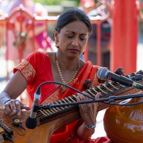Devaki O'Riordan closes her eyes as she plays sitar in a seated position in a sunlit bandstand. She has long dark hair pulled back into a ponytail and wears vivd red traditional dress with adorned with intricate gold patterns.