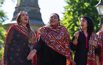 Three members of the Song Geet women's voice choir wearing traditional South Asian dress smile as they sing together in a sunny park.