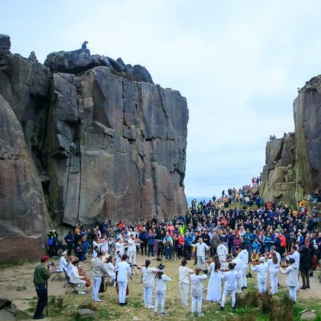 26 members of Paraorchestra dressed in white perform in the centre of a former quarry at dawn. A crowd of several hundred look on, with more spectators watching from the rocks above.