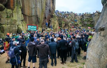 A large crowd gather in the grey dawn light to watch Commoners Choir perform inside the former quarry at Cow and Calf. The are dressed in all black an standing in a loose semi-circle, whilst the audience are perched on rocks an on the lip of the quarry above.