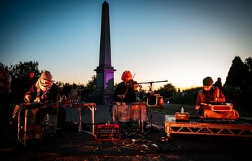 Three musicians perform in Undercliffe Cemetery. From left to right they are using a midi controller, violin, and santoor. Behind them, a large obelisk is silhouetted against the sunset sky.